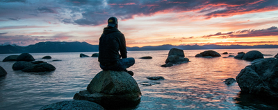 Une personne est assise les jambes crois&eacute;es sur un rocher entour&eacute; d&rsquo;eau. Sa silhouette se d&eacute;coupe sur le coucher de soleil.